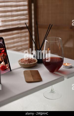 Weißes Holztablett mit einem Glas Wein, Kamm und brennender Kerze auf der Badewanne im Badezimmer Stockfoto