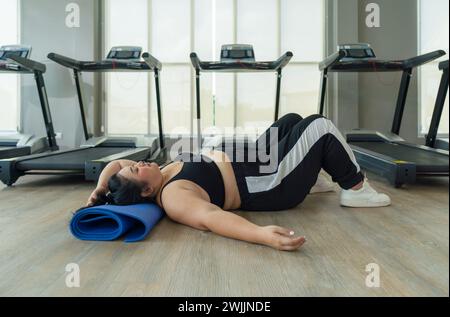 Eine Frau in Übergröße liegt auf dem Boden des Fitnessstudios, ruht oder unmotiviert, umgeben von Trainingsgeräten. Stockfoto