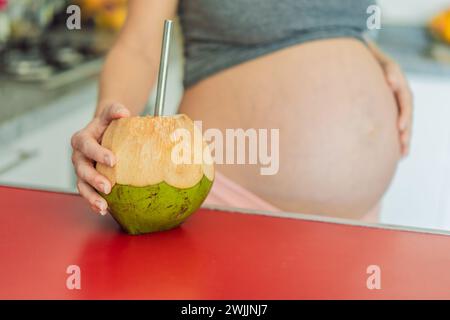 Eine schwangere Frau löscht ihren Schwangerschaftsdurst mit einer erfrischenden Wahl und trinkt freudig Kokoswasser aus einer Kokosnuss in der Küche Stockfoto