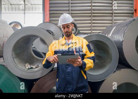 Ein Arbeiter in einem Schutzhelm mit einem Tablet-Computer, der in einem Industrielager oder einer Produktionsstätte vor Stahlspulen steht. Stockfoto