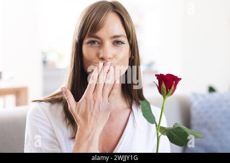 Eine junge kaukasische Frau wirft einen Kuss in die Kamera und hält eine rote Rose in der Hand Stockfoto