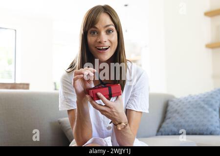 Eine junge Kaukasierin scheint begeistert zu sein, als sie eine kleine rote Geschenkbox öffnet Stockfoto
