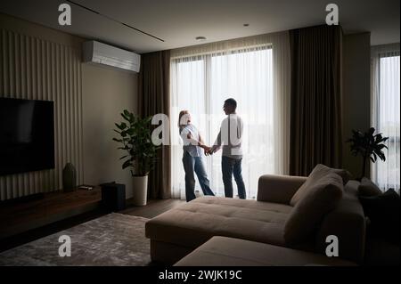 Niedliches Paar, das zu Hause bei natürlichem Sonnenlicht gegenüber dem Fenster steht. Gutaussehender Mann, der mit seiner Freundin Händchen hält. Blick aus der Ferne auf ein Paar in legeren Kleidern. Stockfoto