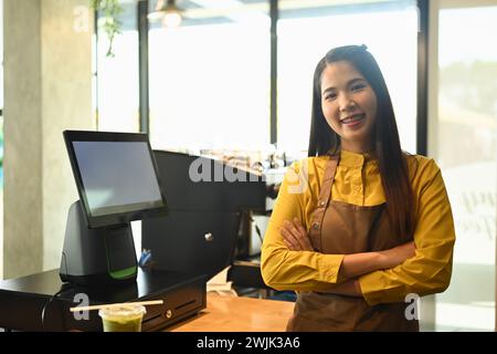 Junger kleiner Coffee Shop Besitzer, der mit überkreuzten Armen hinter dem Tresen mit Kasse steht Stockfoto