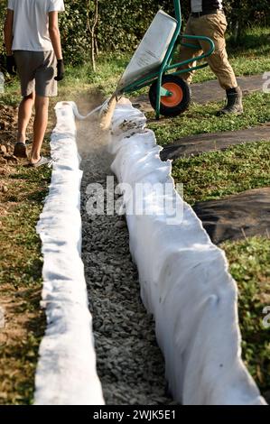 Ein Mann gießt Schutt aus einer Schubkarre in einen Graben. Entwässerungsarbeiten zur Entwässerung des Grundwassers um das Feld herum. Stockfoto