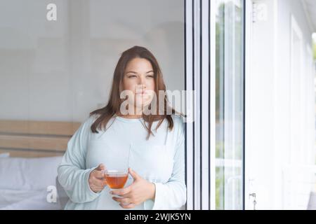 Eine junge Frau in Übergröße genießt einen friedlichen Moment zu Hause und hält einen Drink Stockfoto