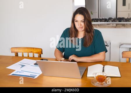 Eine junge Frau mit birassischer Größe arbeitet von zu Hause aus und konzentriert sich unverändert auf ihren Laptop Stockfoto