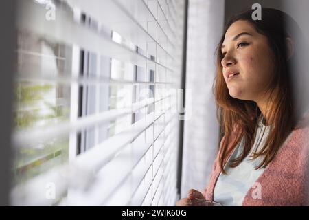 Junge birassische Frau in Übergröße schaut aus einem Fenster zu Hause, mit Kopierraum Stockfoto