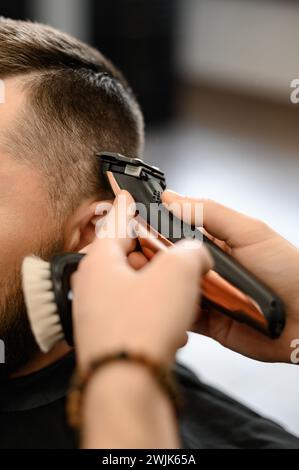 Barber rasiert die Kontur der ovalen Linie mit einem Haarschneider am Kopf des Kunden. Ein Mann mit Bart bekommt einen Haarschnitt in einem Friseurstuhl. Stockfoto