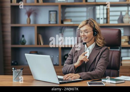 Professionelle Geschäftsfrau mit Headset, die in einem organisierten Heimbüro an ihrem Laptop arbeitet und Kompetenz und Komfort zeigt. Stockfoto