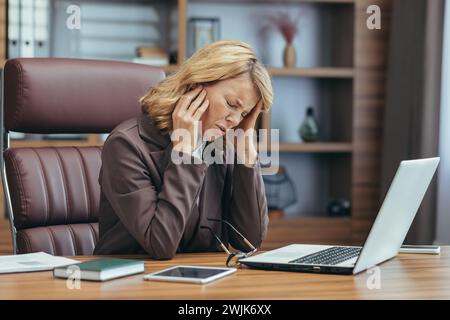 Eine reife Geschäftsfrau fühlt sich überfordert, wenn sie im Home Office an einem Notebook arbeitet und Anzeichen von Stress und Ermüdung zeigt. Stockfoto
