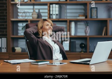Erfahrene Geschäftsfrau, die eine Pause einlegt und sich in einem modernen Heimbüro zufrieden in ihrem Stuhl zurücklehnt. Stockfoto