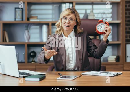 Eine hochrangige Frau in einem Büro hält einen roten Wecker und zeigt Konzepte für Zeitmanagement und Work-Life-Balance. Stockfoto