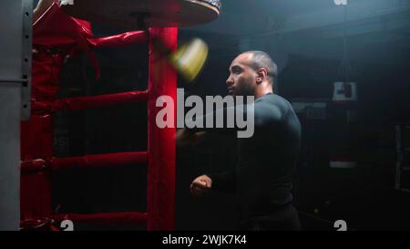 Fokussierter afroamerikanischer Kämpfer trifft beim Workout im dunklen Boxstudio den Boxsack. Sportliche Männer-Übungen vor dem Kampf gegen Wettkämpfe oder Turniere. Boxer bereitet sich auf den Kampf vor und trainiert im Boxstudio Stockfoto