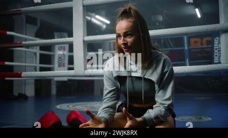 Die Boxerin sitzt in der Nähe des Boxrings und diskutiert die Turnierergebnisse mit dem Interviewer. Athlet spricht nach Kampfmeisterschaft oder Wettkampf im Boxstudio. Boxhandschuhe liegen in der Nähe des Kämpfers. Dolly Shot. Stockfoto