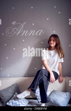 Das schöne Teenager-Mädchen sitzt auf einem Bett in der Nähe der Wand mit ihrem Namen drauf Stockfoto