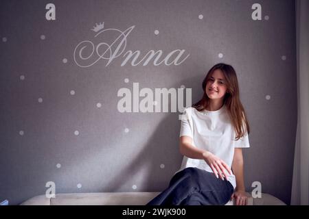 Das schöne Teenager-Mädchen sitzt auf einem Bett in der Nähe der Wand mit ihrem Namen drauf Stockfoto