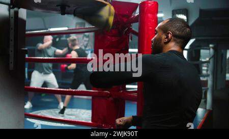 Fokussierter afroamerikanischer Kämpfer trifft beim Workout im dunklen Boxstudio den Boxsack. Sportliche Männer-Übungen vor dem Kampf gegen Wettkämpfe oder Turniere. Boxer bereitet sich auf den Kampf vor und trainiert. Rückansicht. Stockfoto