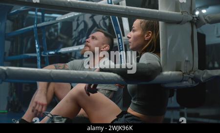 Müde Frau sitzt an der Ringecke, zieht Boxhandschuhe aus und beginnt Wasser zu trinken. Der männliche Trainer setzt sich neben den Boxer und spricht über das Training. Der Athlet bereitet sich auf den Kampf oder Wettkampf im Boxstudio vor. Stockfoto