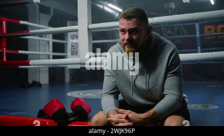 Männlicher Boxer sitzt in der Nähe des Boxrings und spricht mit dem Interviewer über die Turnierergebnisse. Athlet spricht nach Kampfmeisterschaft oder Wettkampf im Boxstudio. Boxhandschuhe liegen in der Nähe des Kämpfers. Dolly Shot. Stockfoto