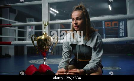 Boxerin sitzt am Boxring und diskutiert mit Interviewer den Sieg im Turnier. Athlet spricht nach Meisterschaft oder Wettkampf im Boxstudio. Eine Tasse Sieger und Boxhandschuhe liegen in der Nähe des Käufers. Stockfoto