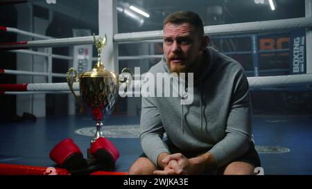 Männlicher Boxer sitzt in der Nähe des Boxrings und spricht mit dem Interviewer über den Sieg im Turnier. Athlet spricht nach Meisterschaft oder Wettkampf im Boxstudio. Eine Tasse Sieger und Boxhandschuhe liegen in der Nähe des Käufers. Stockfoto