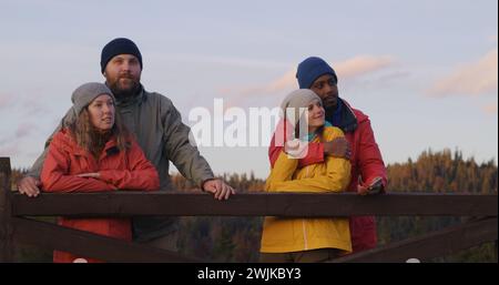 Multiethnische Paare stehen in der Nähe eines Holzzauns, sehen Sie den wunderschönen Sonnenuntergang und genießen die Natur während des Ausflugs. Gruppe von Touristen oder Wanderfreunden ruhen sich nach der Wanderung in den Bergen aus. Outdoor-Enthusiasten. Zeitlupe. Stockfoto