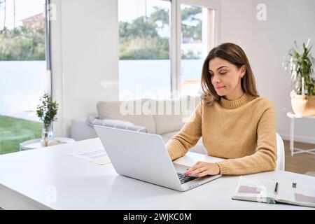 Lächelnde reife Frau, die einen Laptop benutzt und am Tisch im Wohnzimmer sitzt. Stockfoto
