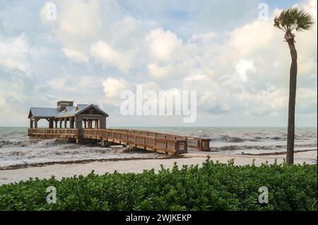 Ein hölzerner Pier mit einem überdachten Teil am Ende, der in ein raues Meer reicht, mit tropischem Laub im Vordergrund Stockfoto