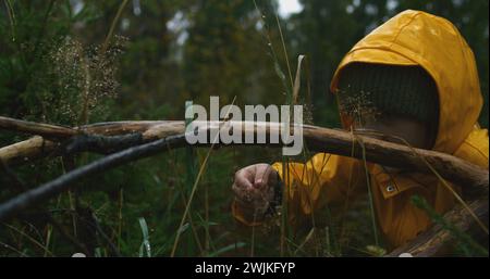 Filmische Aufnahme eines Mädchens mit gelbem Regenmantel, das in den regnerischen Wäldern spaziert, während es in den Bergen wandert. Junge Touristen berühren Regentropfen auf nassen Baumzweigen im Wald. Outdoor-Erkundung. Zeitlupe. Stockfoto