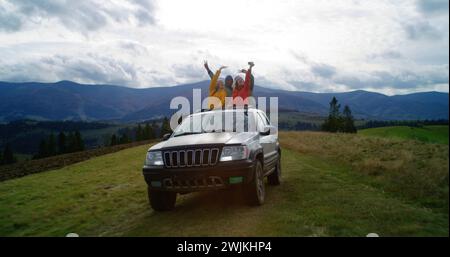 Eine Gruppe multiethnischer Touristen reitet gemeinsam in den Bergen auf Urlaub. Wanderer stehen durch das Sonnendach des Autos, lachen und winken auf dem Roadtrip mit den Händen. Genieße das Fahren in einer Autoluke. Outdoor-Enthusiasten. Stockfoto