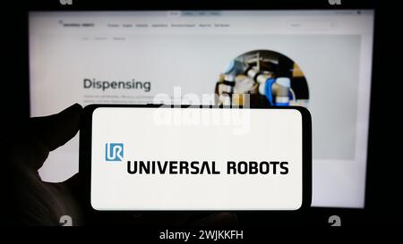 Person, die ein Mobiltelefon mit dem Logo des dänischen Cobot-Unternehmens Universal Robots hält, WIE vor der Business-Webseite. Schwerpunkt auf dem Display des Telefons. Stockfoto