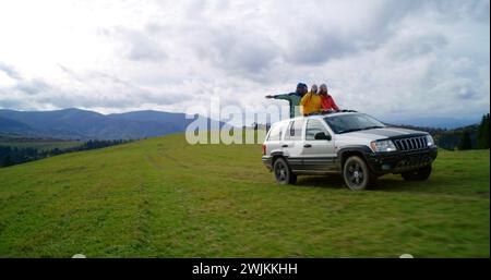 Eine Gruppe multiethnischer Touristen reitet gemeinsam in den Bergen auf Urlaub. Wanderer stehen durch das Sonnendach des Autos, lachen und winken auf dem Roadtrip mit den Händen. Genieße das Fahren in einer Autoluke. Outdoor-Enthusiasten. Stockfoto