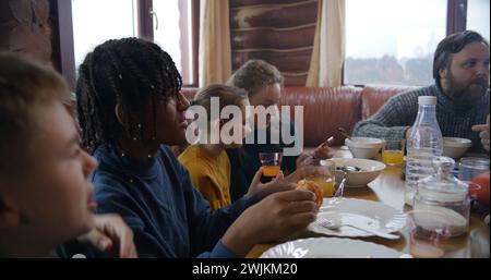 Große, vielfältige Familie sitzt am Tisch, isst, trinkt, redet. Yong Girl mit Mom Dip Pancakes in Kondensmilch. Eine Gruppe von Freunden versammelte sich im Ferienhaus. Touristen ruhen sich während des Ausflugs in einem Holzhaus aus. Stockfoto