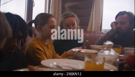 Große, vielfältige Familie sitzt am Tisch, isst, trinkt, redet. Yong Girl mit Mom Dip Pancakes in Kondensmilch. Eine Gruppe von Freunden versammelte sich im Ferienhaus. Touristen ruhen sich während des Ausflugs in einem Holzhaus aus. Stockfoto