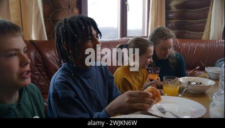 Große, vielfältige Familie sitzt am Tisch, isst, trinkt, redet. Yong Girl mit Mom Dip Pancakes in Kondensmilch. Eine Gruppe von Freunden versammelte sich im Ferienhaus. Touristen ruhen sich während des Ausflugs in einem Holzhaus aus. Stockfoto