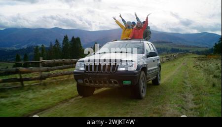 Eine Gruppe multiethnischer Touristen reitet gemeinsam in den Bergen auf Urlaub. Wanderer stehen durch das Sonnendach des Autos, lachen und winken auf dem Roadtrip mit den Händen. Genieße das Fahren in einer Autoluke. Outdoor-Enthusiasten. Stockfoto