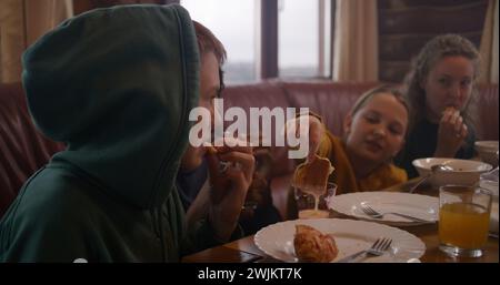 Große, vielfältige Familie sitzt am Tisch, isst, trinkt, redet. Yong Girl mit Mom Dip Pancakes in Kondensmilch. Eine Gruppe von Freunden versammelte sich im Ferienhaus. Touristen ruhen sich während des Ausflugs in einem Holzhaus aus. Stockfoto