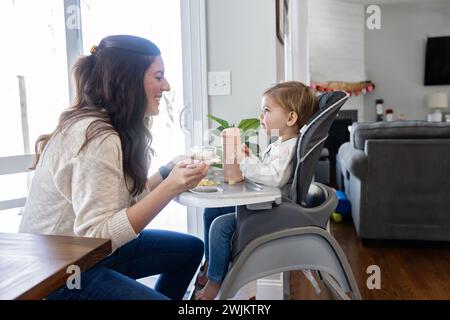 Die Mutter lächelt die Tochter des Kleinkindes an, während sie ihr Mittagessen füttert Stockfoto