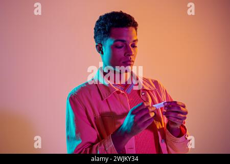 afroamerikanischer Mann, der Zigarette für das Rauchen auf buntem Hintergrund mit blauer Neonbeleuchtung rollt Stockfoto