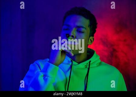 Ernster afroamerikanischer junger Mann im Hoodie, der Zigarre auf dunkelblauem Hintergrund mit rotem Licht raucht Stockfoto