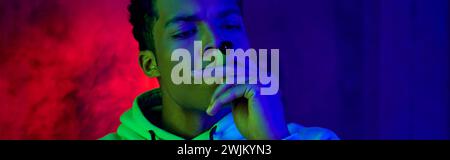 Banner des afroamerikanischen jungen Mannes im Hoodie, der Zigarre auf dunkelblauem Hintergrund mit rotem Licht raucht Stockfoto