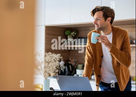 Nachdenklicher junger Mann, der eine Tasse Kaffee trinkt, während er in der Küche steht Stockfoto