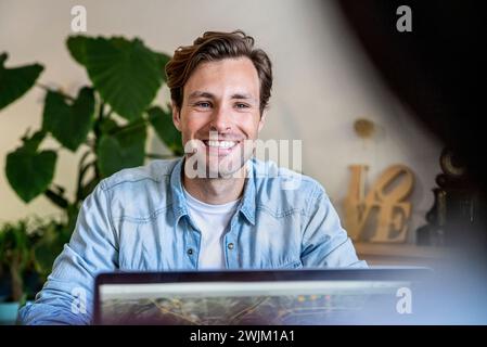 Fröhlicher männlicher Grafikdesigner, der am Schreibtisch sitzt und einen Laptop benutzt Stockfoto