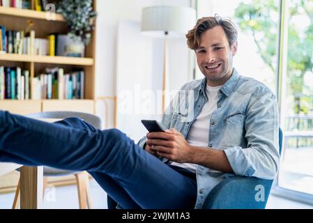 Geschäftsmann, der das Smartphone benutzt, während er auf dem Stuhl sitzt und die Füße auf dem Tisch liegen Stockfoto