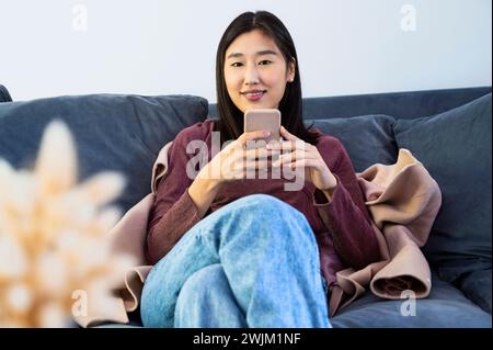 Junge Erwachsene Frau, die auf die Kamera blickt, während sie ihr Smartphone benutzt Stockfoto