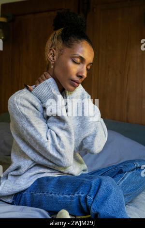 Eine gestresste junge Erwachsene Frau, die ihren Hals berührt, während sie auf dem Bett sitzt Stockfoto