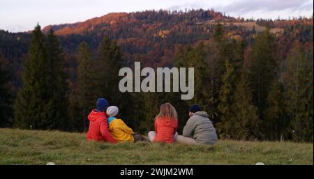 Multiethnische Paare sitzen auf dem Gras auf der Spitze des Hügels, betrachten den Sonnenuntergang und die atemberaubende Bergkulisse. Eine Gruppe von Touristen oder Wanderfreunden genießt die wunderschöne Natur während der Reise. Outdoor-Enthusiasten. Stockfoto