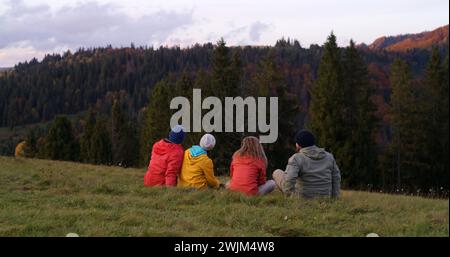 Multiethnische Paare sitzen auf dem Gras auf der Spitze des Hügels, betrachten den Sonnenuntergang und die atemberaubende Bergkulisse. Eine Gruppe von Touristen oder Wanderfreunden genießt die wunderschöne Natur während der Reise. Outdoor-Enthusiasten. Stockfoto