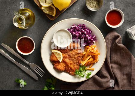 Schnitzel mit Kartoffelfrites, Rotkohlsalat und Sauce auf weißem Teller auf dunklem Steinhintergrund. Draufsicht, flach Stockfoto